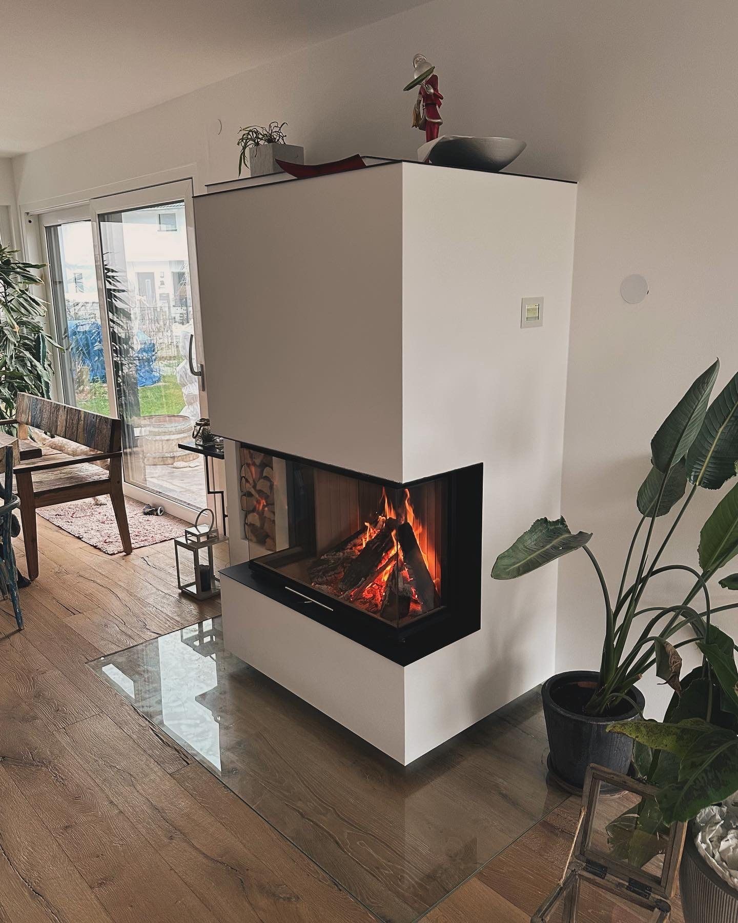Weißer Eckkamin mit Feuer im Wohnraum, davor eine Glasbodenplatte auf Holzdielen