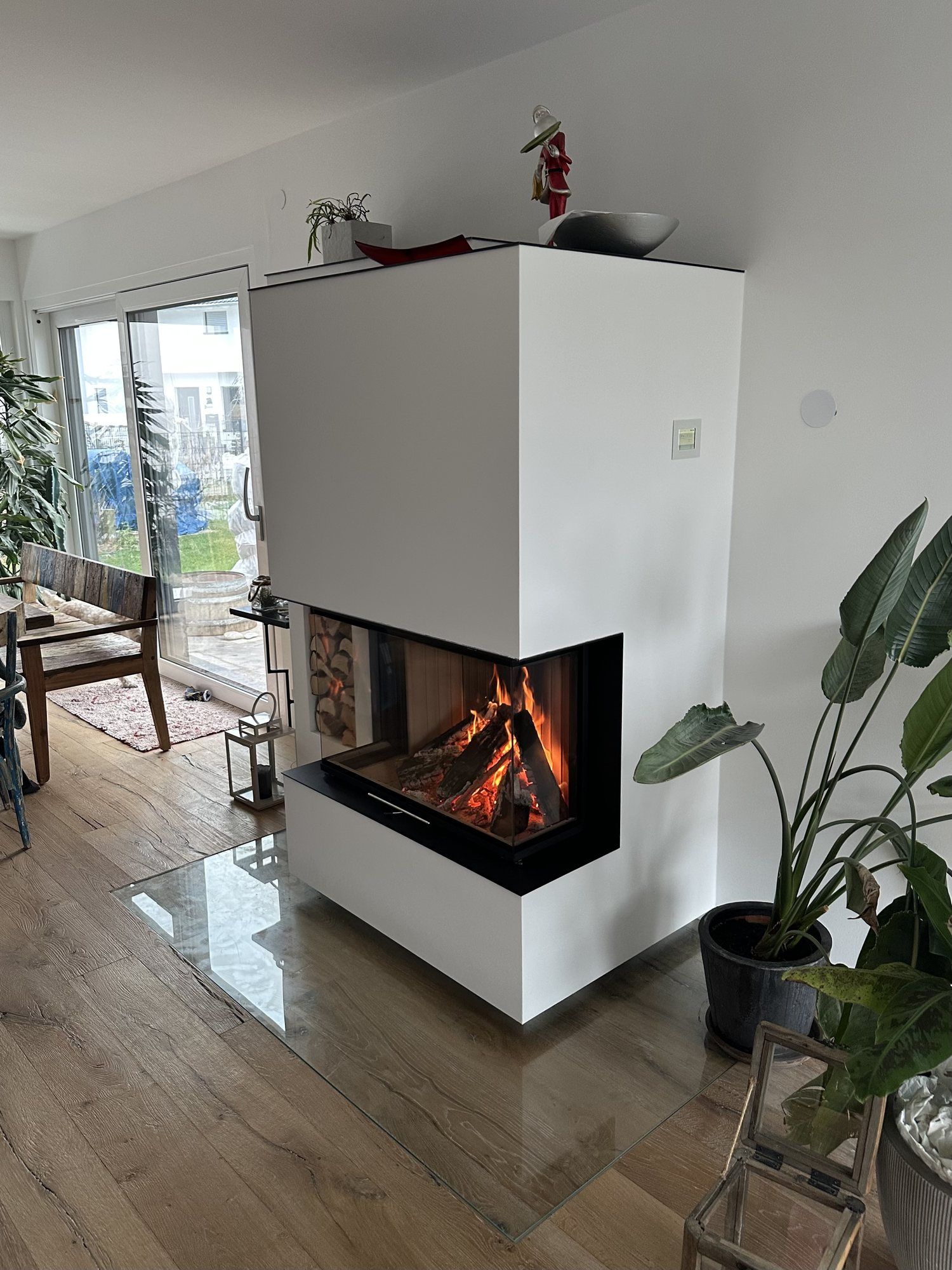 Weiß verputzter Heizkamin mit Eck-Sichtscheibe und Feuer in einem hellen Wohnraum mit Holzboden