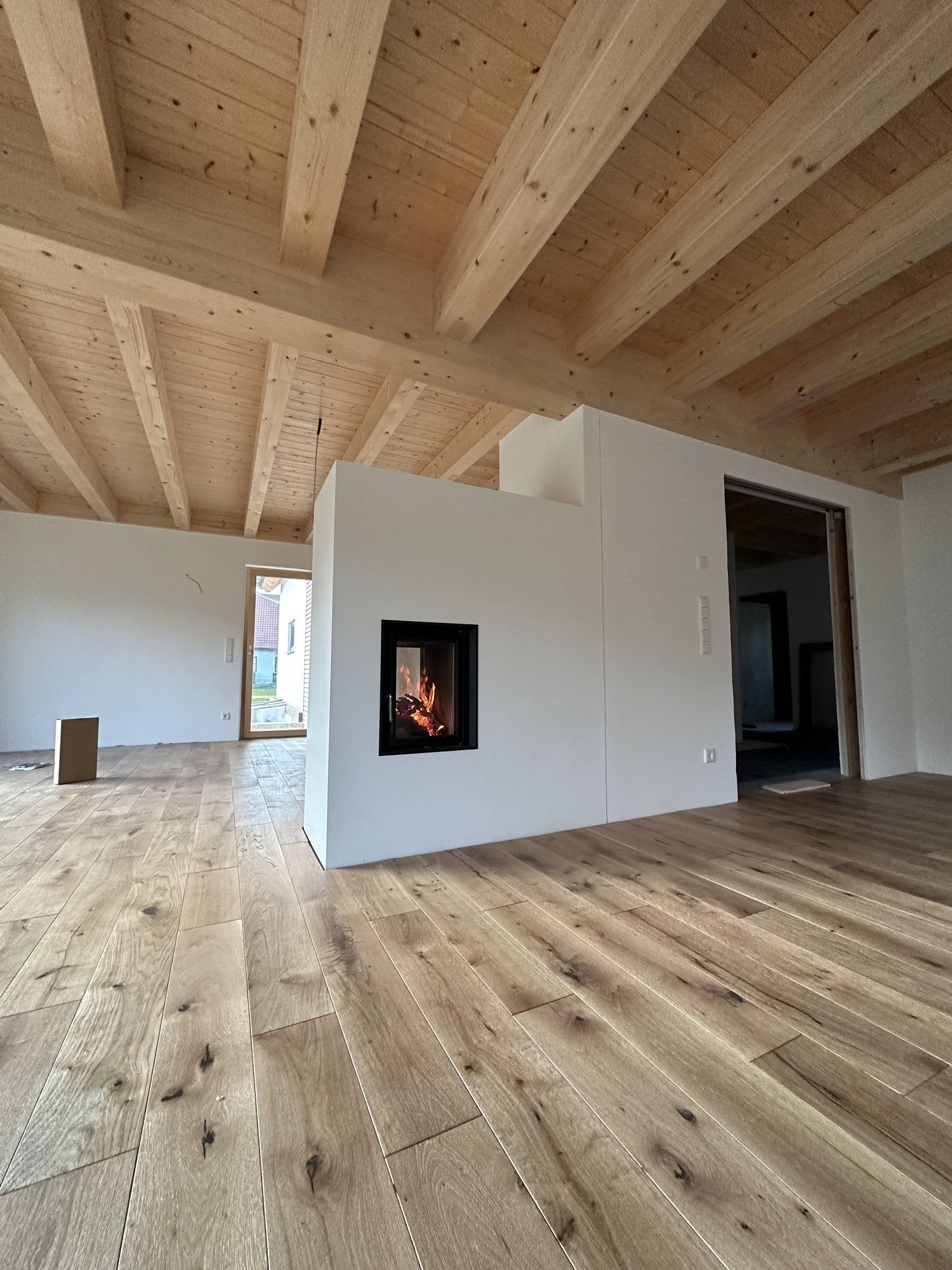 Kamin als weißer Raumteiler in einem offenen Wohnraum mit sichtbarer Holzbalkendecke und Eichendielen